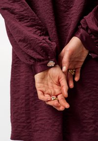 Dieprode tekstuur jurk met opgestoken mouwen, voorzien van een zilveren decoratieve knop op de manchet. Handen met ringen zichtbaar op de voorgrond.
