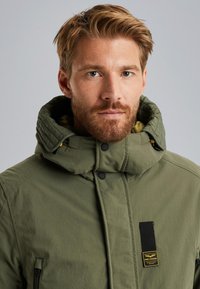 Groene geïsoleerde jas met een hoge kraag en aangehechte capuchon, voorzien van een patchlogo, ritszakken en een gestructureerde voering.
