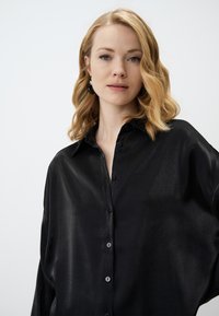 Black button-up shirt made of a smooth, shiny fabric. Features a spread collar and loose fit with button details along the front.