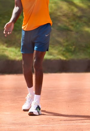 Oranges Sportoberteil, marineblaue Shorts, weiße Sneakers mit grünen Akzenten. Steht auf einem roten Tennisplatz, Füße in Bewegung.