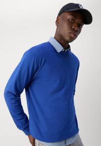 Suéter azul de manga larga con cuello redondo, que presenta un collar de camisa a cuadros debajo. Textura suave y un sutil detalle de logo.