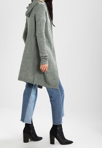 Kvinna som går klädd i en lång grå stickad kardigan med huva, tvåtonade beskjutna jeans och svarta klackiga ankelstövlar.