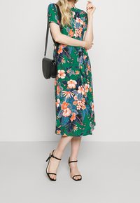 Zielona sukienka w kwiatowy wzór z krótkimi rękawami, z pomarańczowymi i różowymi kwiatami. W komplecie z czarnymi sandałami na obcasie i czarną torebką typu crossbody.