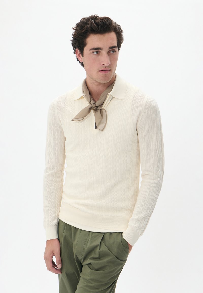 Pulóver de punto acanalado color crema con cuello, combinado con una bufanda beige. Pantalones verdes oliva de corte holgado. Textura suave, diseño minimalista.