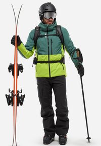 La tenue de ski comprend une veste verte et lime à finition texturée, un pantalon noir, des gants noirs et des skis orange, accompagnés d'un bâton de ski.