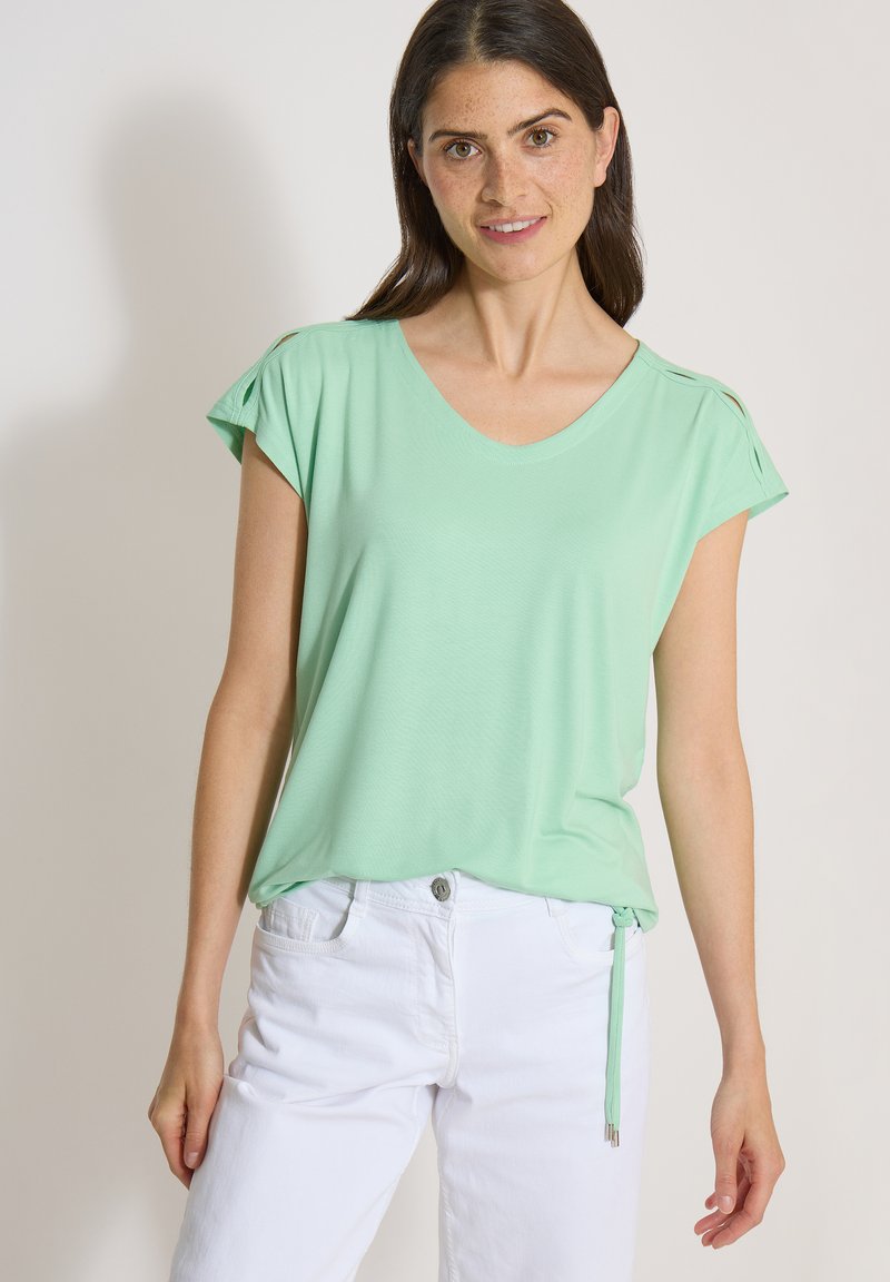 Jeune femme portant un haut vert menthe à manches courtes avec un pantalon blanc, debout devant un fond clair uni.
