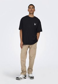 Man staat in een zwart oversized T-shirt met een klein vlinderlogo, beige cargobroeken en zwart-witte sneakers op een effen achtergrond.