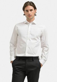 Vit långärmad skjorta med standardkrage, subtil textur, knapplinkning framtill och rundad nederkant, kombinerad med svarta byxor.