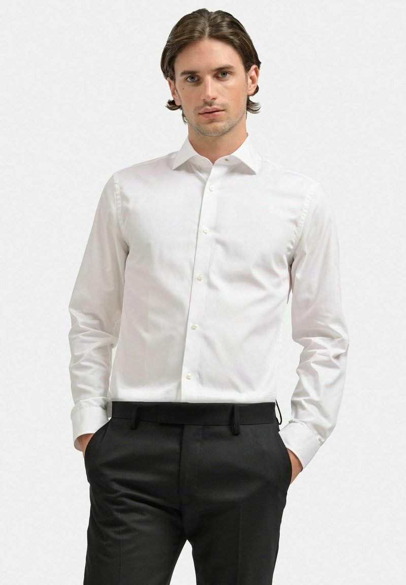 Vit långärmad skjorta med standardkrage, subtil textur, knapplinkning framtill och rundad nederkant, kombinerad med svarta byxor.