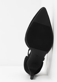 Pompe en suede noir avec bout pointu et talon aiguille. Semelle en caoutchouc texturé avec des rainures verticales. Sangle ajustable à la cheville.