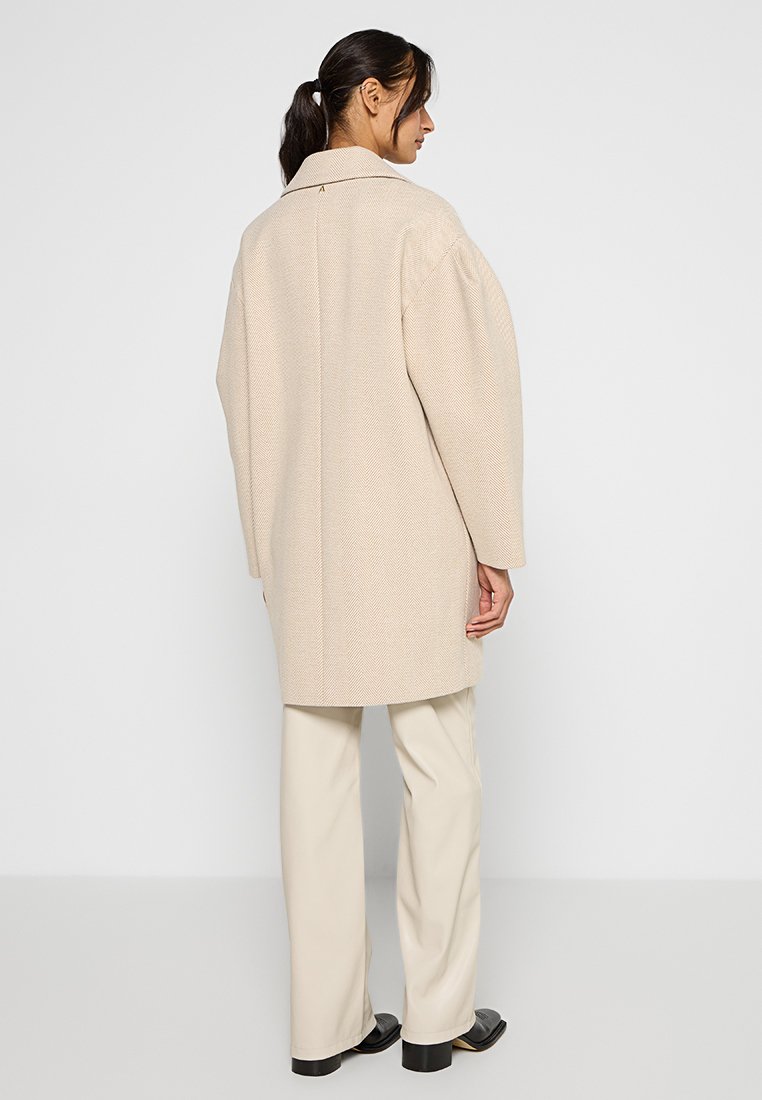 Beżowy płaszcz w jodełkę z kołnierzem, długimi rękawami i swobodnym krojem, zestawiony z kremowymi spodniami i czarnymi butami. Widok z tyłu.