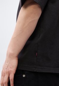 Κοντινή λήψη του αριστερού βραχίονα και του χεριού ενός ατόμου που ακουμπούν δίπλα στην τσέπη του μαύρου πουκαμίσου και μαύρου παντελονιού με μια μικρή κόκκινη ετικέτα.