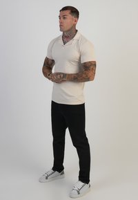 Beige polo shirt voor heren met korte mouwen, gecombineerd met zwarte broeken en witte sneakers met grijze accenten. Model staat met zijn handen gevouwen.