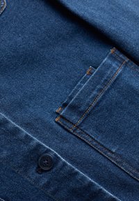 Tmavě modrý denim s kontrastním oranžovým prošíváním, s knoflíkovým zapínáním a malou přední kapsou. Hladká textura s pevným designem.