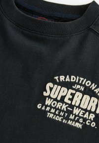 Sweatshirt negru cu guler rotund din material elastic, având text alb brodat: "TRADITIONAL JPN SUPERDRY WORK-WEAR" și elemente decorative.