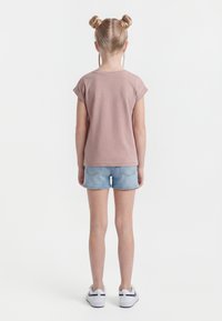 Dieťa s blond vlasmi v dvoch bočných drdoloch, oblečené v fialovo-ružovom tričku s krátkym rukávom, svetlých riflových šortkách a bielych teniskách, stojí chrbtom.