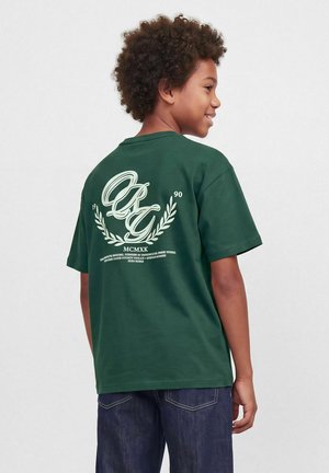 Un enfant souriant regarde par-dessus son épaule, montrant un T-shirt vert foncé avec une inscription en écriture cursive couleur crème et un motif de laurier, porté avec un jean.