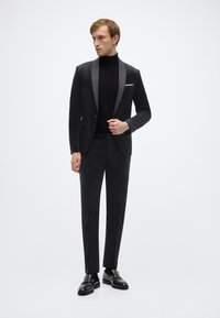 Schwarzer Samtanzug mit Satinrevers, angepasster Silhouette, darunter ein Rollkragenpullover und glänzende schwarze Loafers. Einfaches, elegantes Design, keine sichtbaren Muster.