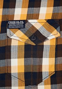 Rutiga skjortfickor med gula, bruna, vita och marinblå rutor, fästa med en svart knapp, som har en etikett för industriellt arbetskläder från Crude Oil Co.