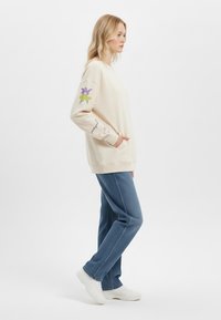 Felpa crema con fiore viola ricamato e dettagli verdi sul maniche sinistra, abbinata a pantaloni in denim blu e sneakers bianche.