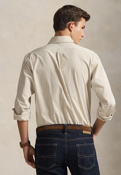 Smėlio ir baltos spalvos dryžuoti marškiniai su sagomis, rankovės užsukti, dėvimi su tamsiai mėlynais džinsais ir rudos spalvos diržu, žiūrint iš nugaros.