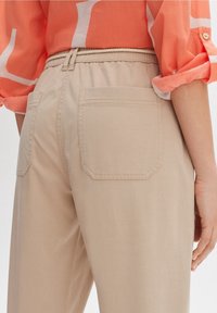 Pantalon beige avec une taille élastique et deux poches arrière. Texture lisse et coupe ajustée, associé avec une chemise orange vif.