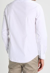 Man in een lange mouw witte overhemd en beige broek, gezien van achteren tegen een effen lichte achtergrond.