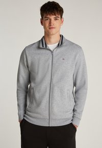 Joven con chaqueta gris claro de cremallera y cuello a rayas, y pantalones negros, de pie con las manos en los bolsillos frente a un fondo liso.