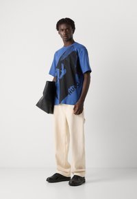 Zila un melna grafiskā T-kreklā ar tekstu un dizaina elementiem; bēšas bikses; melna soma; melni sporta apavi. Vienkāršs, modernos sportiskā stilā.