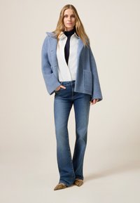 Chaqueta de punto azul claro con mangas anchas y bolsillo, llevada sobre una camisa blanca y una bufanda oscura, combinada con jeans azules de campana y zapatos con estampado.