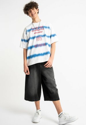 Nastolatek uśmiecha się, ma na sobie białą i niebieską pasiastą oversize koszulkę z napisem, czarne szerokie krótkie spodenki do kolan i białe trampki.