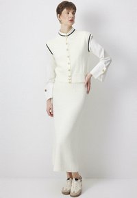 Witte, gebreide tweedelige set bestaande uit een mouwloze top met gouden knopen en zwarte accenten, gecombineerd met een lange, strakke rok. Gedragen met beige sneakers.
