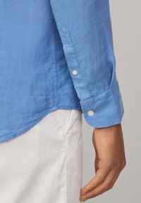 Světle modrá lněná košile s hladkou strukturou; má manžety na knoflíky a zakřivený lem. Viditelné švy podél okrajů.