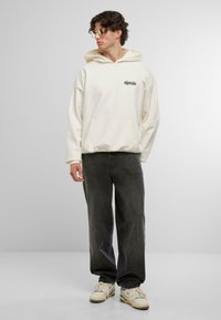 Felpa oversize color crema con tasca frontale e testo nero, abbinata a jeans scuri e sneakers bianche. Il modello indossa occhiali da sole trasparenti.