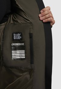 Olive green jacket with shiny, smooth fabric. Features a zippered pocket, tags with black and white text, and a textured black accent.