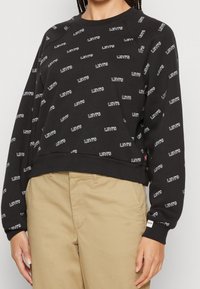 Felpa nera con logo "Levi's" bianco stampato su tutta la superficie, caratterizzata da un collo rotondo e polsini a costine, abbinata a pantaloni beige.