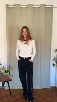 Hosszú ujjú fehér felső kék logóval, fekete magas derekú farmerrel párosítva, világosszürke függöny és zöld növény előtt állva.