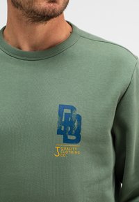 Groene sweatshirt met een textuur, ronde hals en opvallende grafische elementen: een blauw logo en oranje tekst op de linkerkant van de borst.