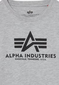 Pelēka kokvilnas T-krekls ar melnu logo, uz kura redzams teksts "ALPHA INDUSTRIES" un stilizēts simbols. Apaļš kakla izgriezums, īsas piedurknes.