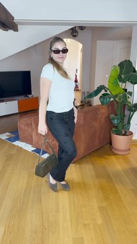 Jeans de ganga pretos com bordados florais, t-shirt branca de mangas curtas, sapatos rasos cinzentos e uma bolsa verde-escura. Sofá de veludo castanho ao fundo.