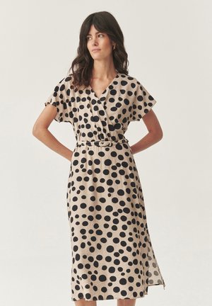 Femme portant une robe midi beige avec de grands pois noirs, à manches courtes, en V, et une fine ceinture, se tenant les mains sur les hanches.