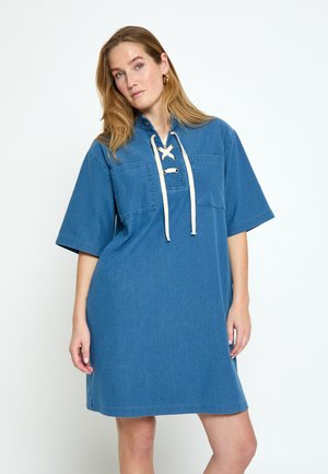 Žena v voľných modrých rifľových šatách s krátkymi rukávmi a predným šnurovaním na výstrihu, stojaca pred jednoduchým bielym pozadím.