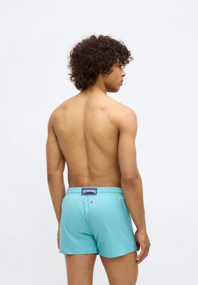 Vīrietis ar cirtainiem matiem stāv profilā, uzvilcis tirkīza peldbikses ar Vilebrequin logo uz aizmugurējā jostas posma.