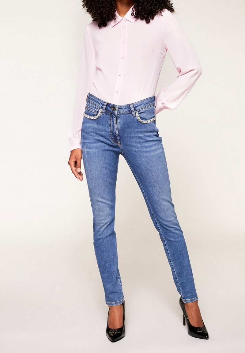 Blusa de color rosa claro con botones y mangas largas, combinada con jeans skinny de mezclilla azul con bolsillos adornados, complementados con tacones negros.