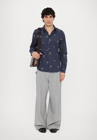 Camicia blu con bottoni decorata con una stampa di palme verdi, abbinata a pantaloni grigi chiari a gamba larga. Il modello tiene una borsa a tracolla marrone e indossa scarpe nere.