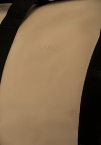 Borsa impermeabile beige con finitura opaca, dotata di cinturini neri e logo "RAINS" in rilievo. Texture liscia, design minimalista.