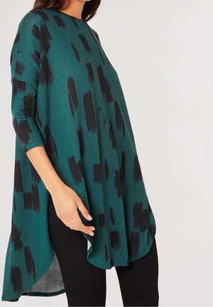 Femme portant une tunique verte ample et asymétrique avec des motifs abstraits noirs en forme de coups de pinceau et des manches trois-quarts, associée à des leggings noirs.