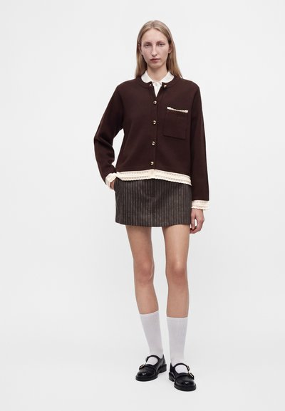Femme portant un cardigan marron boutonné avec une bordure blanche, une mini-jupe à fines rayures, des chaussettes blanches montantes et des chaussures Mary Jane noires, debout sur un fond blanc.