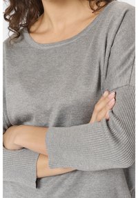 Femme aux bras croisés portant un pull en maille gris clair avec des manches longues côtelées et un col rond.