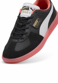 Fekete sneaker velúr és bőr anyagokkal, fehér és rózsaszín kiegészítőkkel, arany márkázással a nyelvén, és áttetsző rózsaszín gumitalppal.
